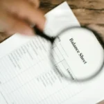 Neraca keuangan sedang diperiksa dengan kaca pembesar di atas meja kayu